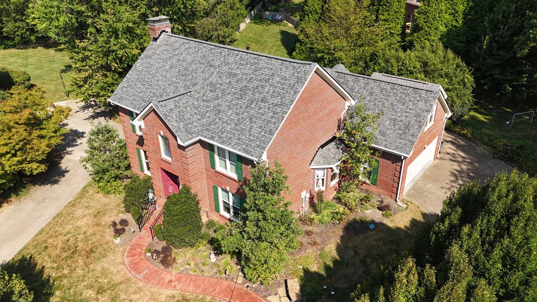 Gray shingle roof on a red brick home with green shutters and a brick walk to the front door, seen from the air