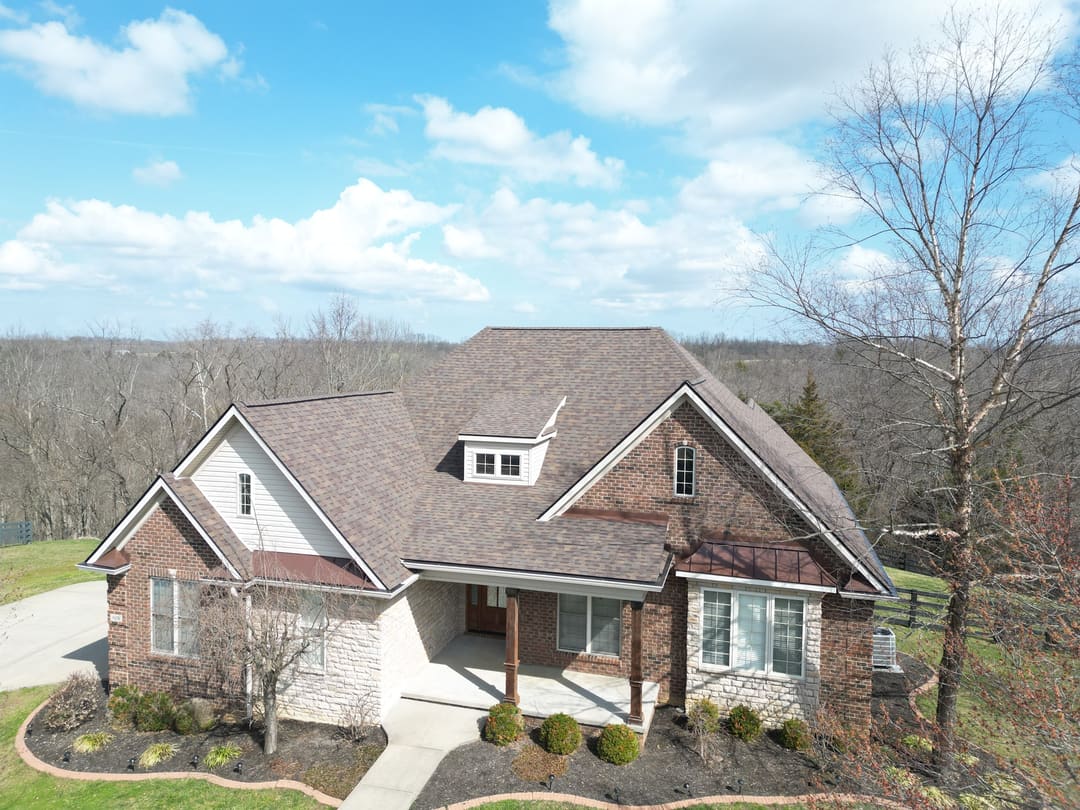 Front of a brick and stone home with a brown shingle roof, white dormer and copper porch roof