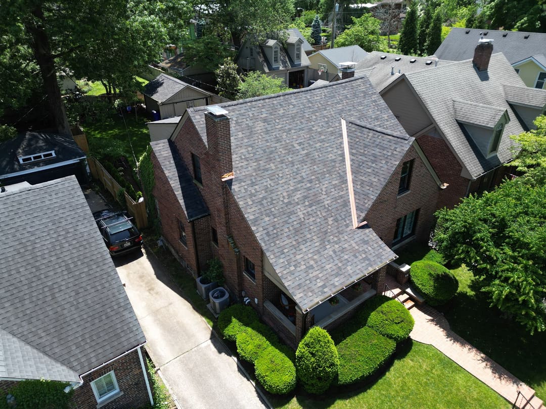 Steep shingle roof on a brick home with a tall chimney, seen from above among trees
