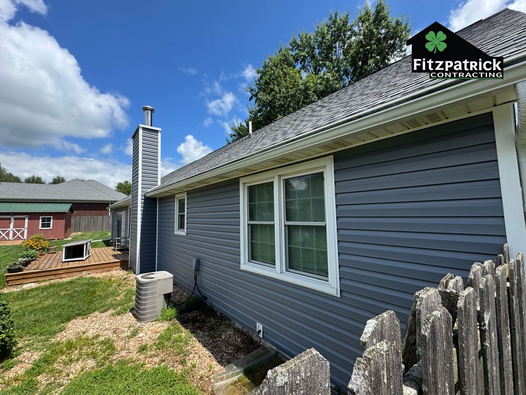 Gray vinyl siding along the side wall of a home, with a wrapped chimney chase and a window in white trim