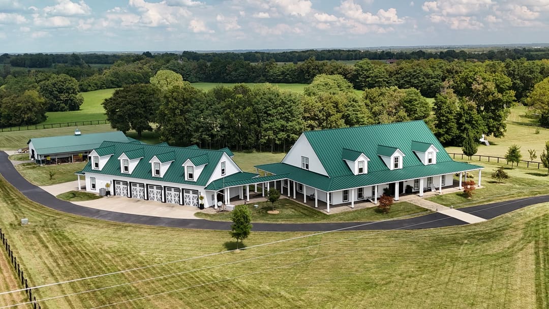 Green standing seam metal roofs on a long white farmhouse and its matching wing, seen from the air over open pasture