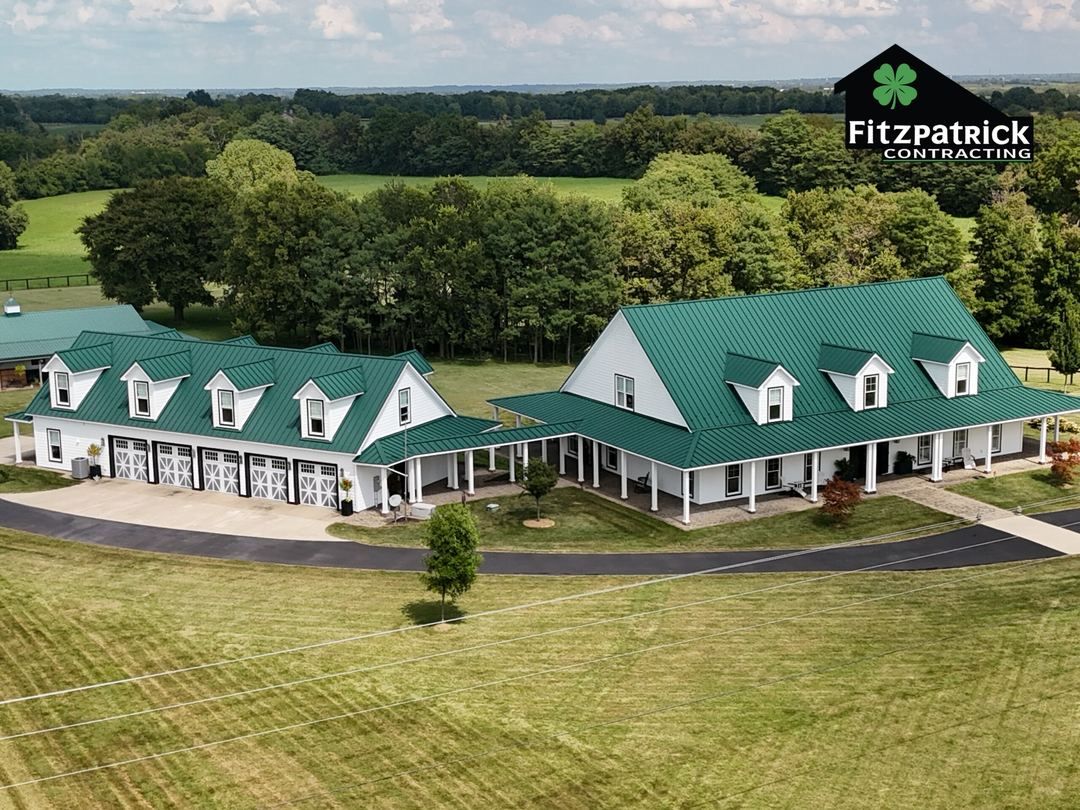 Green standing seam metal roofs on a white farmhouse and its garage wing, with the paved drive and open pasture around them