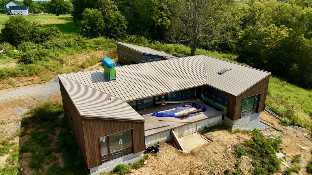 Tan standing seam metal roof on a modern L shaped home with dark wood siding, seen from the air