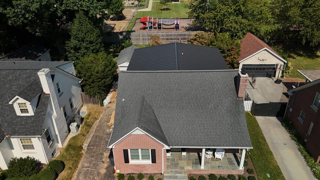 Black standing seam roof on the rear addition of a brick home with a gray shingle roof in front, seen from above