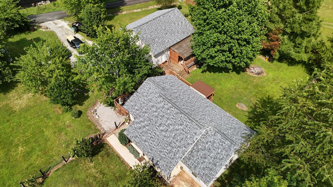 Gray shingle roofs on a ranch home and the outbuilding behind it, with a gravel drive, seen from above