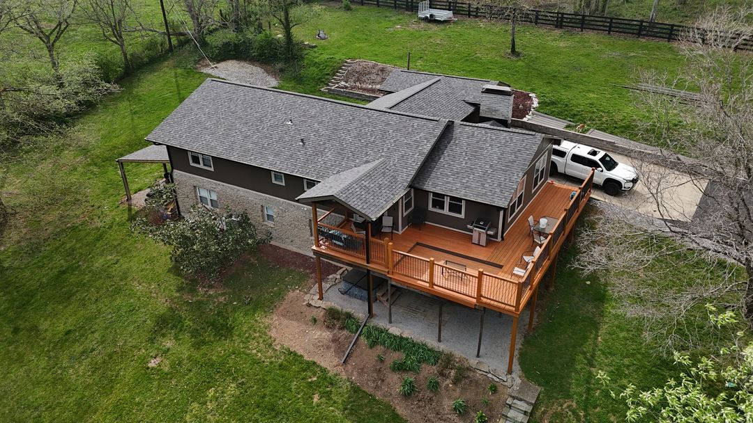 Gray shingle roof on a brown ranch home with a large wood deck and a white truck in the drive, seen from the air