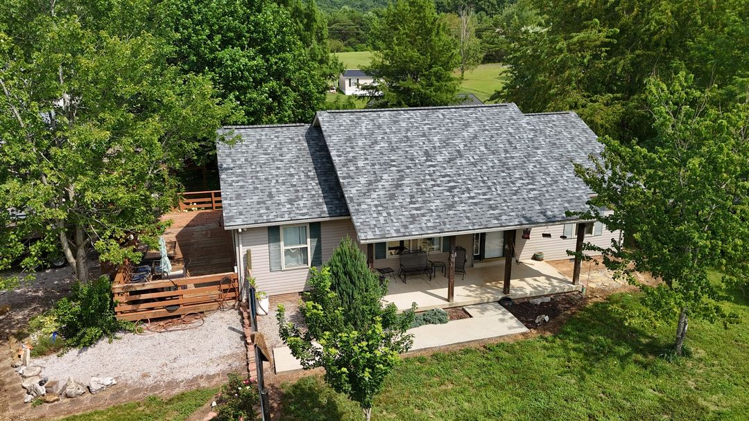 Gray shingle roof on a cream ranch home with a covered porch, a wood deck and a gravel walk, seen from the air