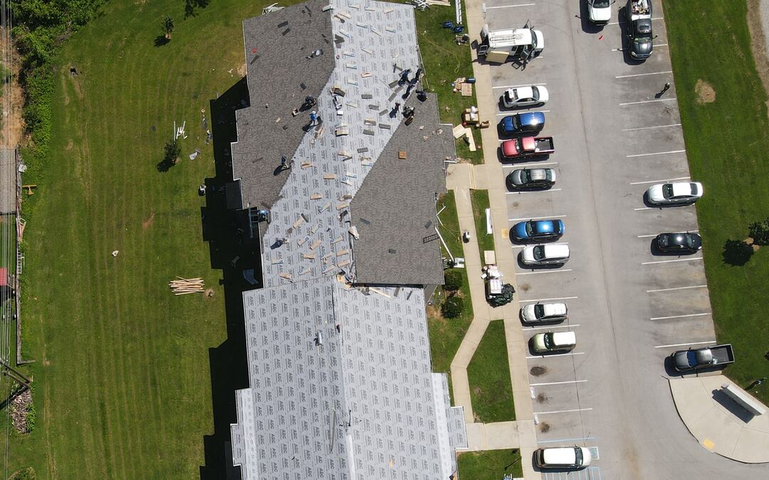 Overhead view of a long apartment roof partway through a reroof, with underlayment down, shingles going on and a crew working along the ridge